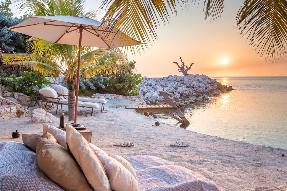 Beach scene at sunset, featuring lounge chairs, a sun umbrella, palm trees, and ocean.