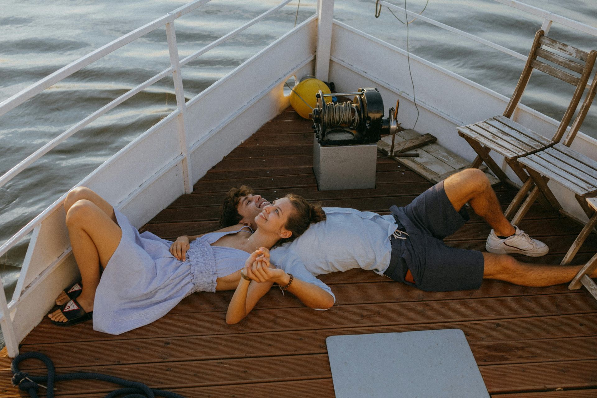 Couple lying on a boat deck, smiling. Woman in blue dress, man in shorts, holding hands. Boat on water.