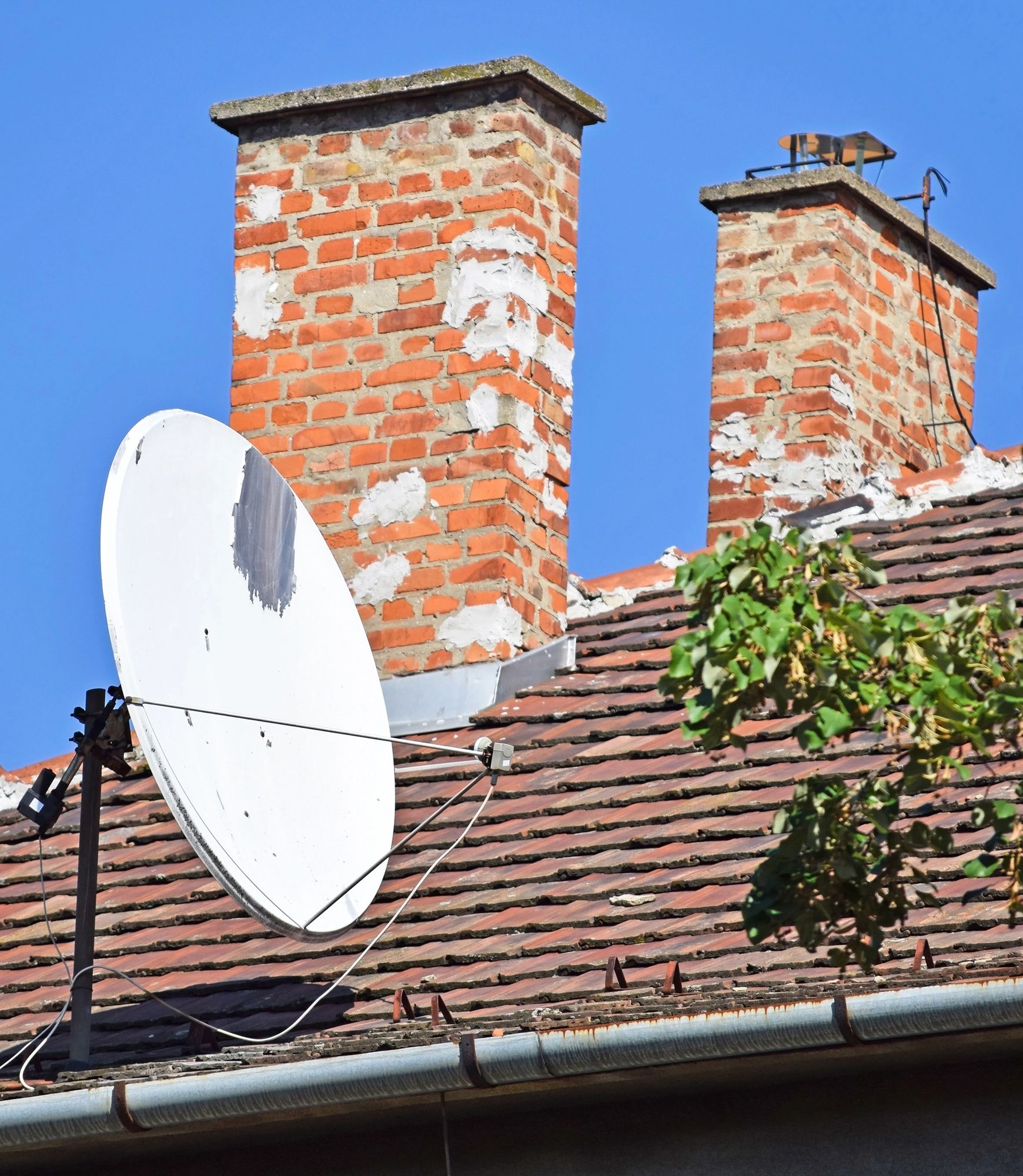 Un'antenna parabolica montata su un tetto con due camini in mattoni consumati dal tempo, stagliarsi contro un cielo azzurro e limpido.