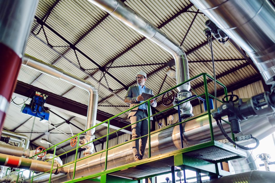 Un ingegnere con l'elmetto cammina lungo una piattaforma metallica rialzata all'interno di un grande stabilimento industriale pieno di tubature.