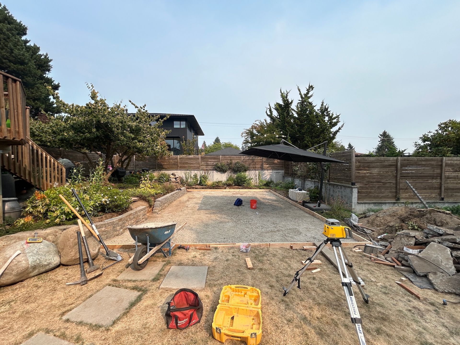Backyard construction site with gravel, tools, wheelbarrow, and fence under a partly cloudy sky