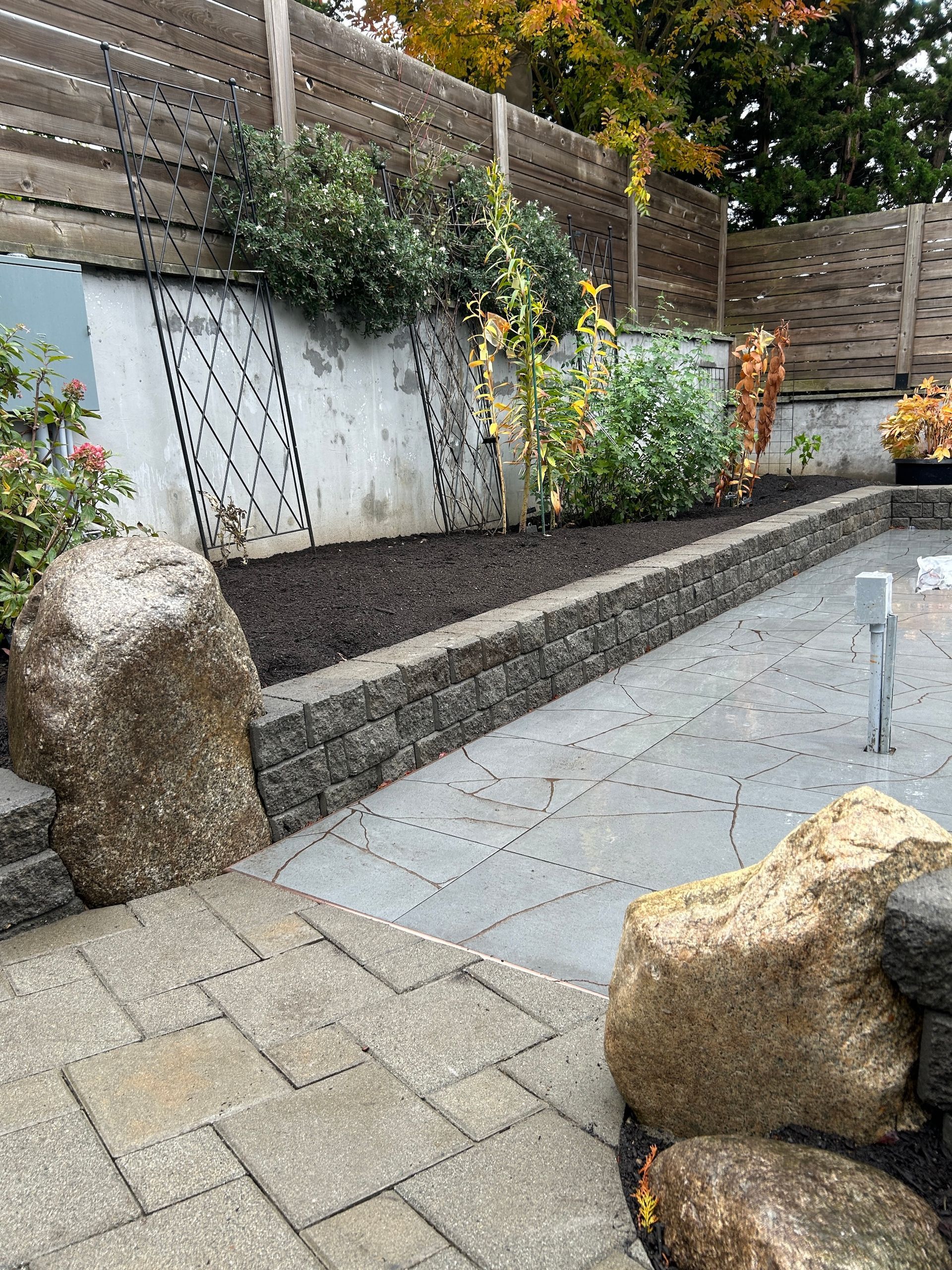 Stone garden patio with sloped planter wall, large rocks, and paved walkway leading to a seating area