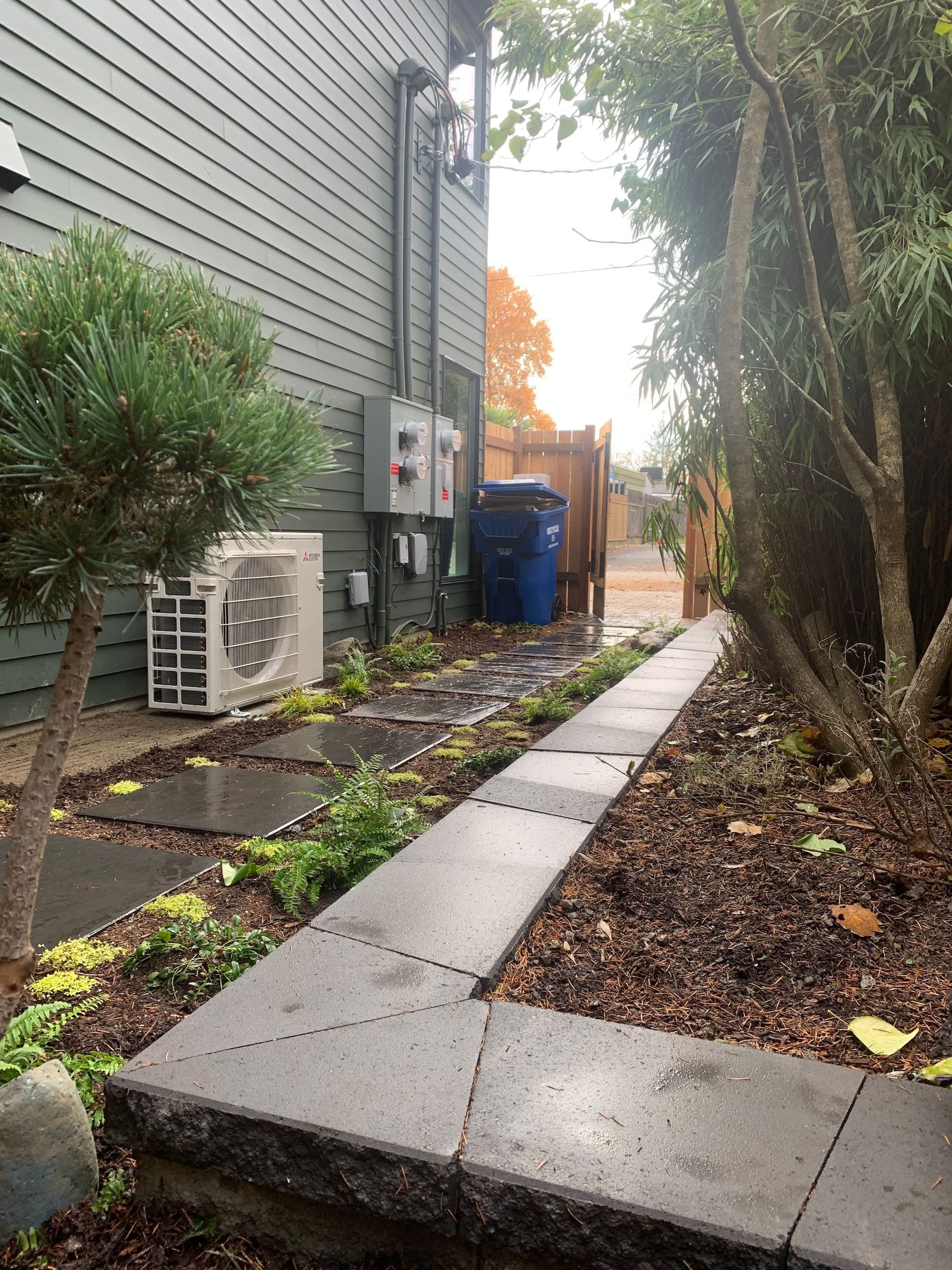 Narrow wet side yard with concrete walkway, house siding, utility units, blue bin, and trees on the right