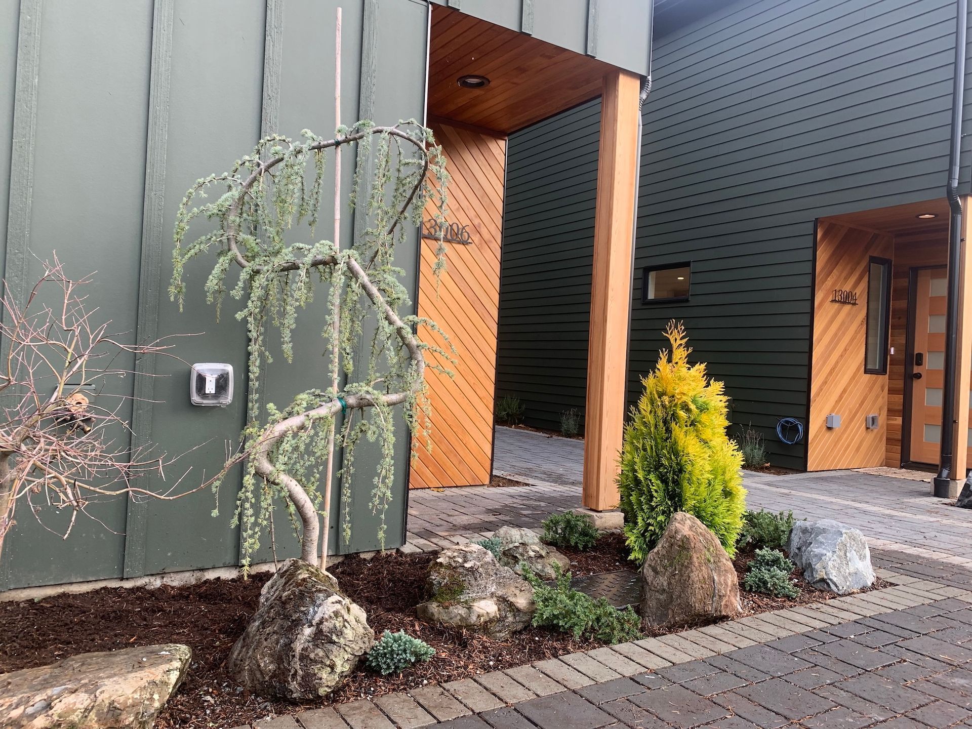 Modern house entryway with dark siding, brick walkway, and a small landscaped garden bed