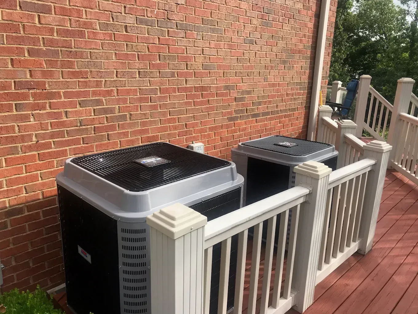 Two air conditioners are sitting on a deck next to a brick wall.