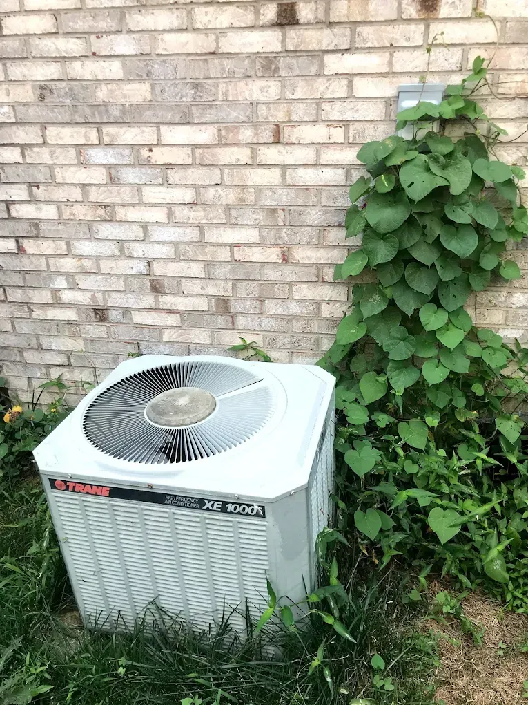 A white air conditioner is sitting in the grass next to a brick wall.