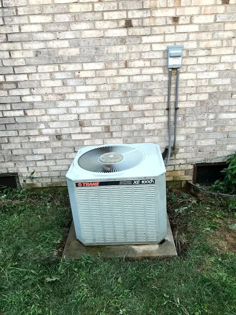 A white air conditioner is sitting in the grass in front of a brick wall.