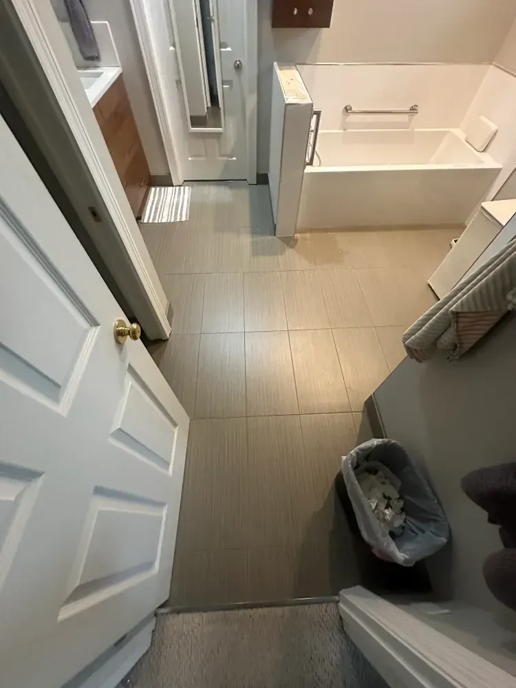 Bathroom hallway with gray tile floor, white door, and bathtub. A trash can and paper towels are visible.