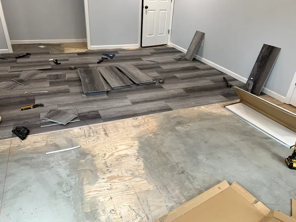 Room with grey flooring partially installed, tools and materials scattered on the concrete floor.