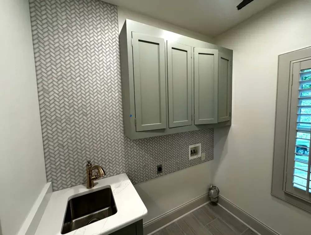 Laundry room with gray cabinets, sink, patterned backsplash, and window with shutters.