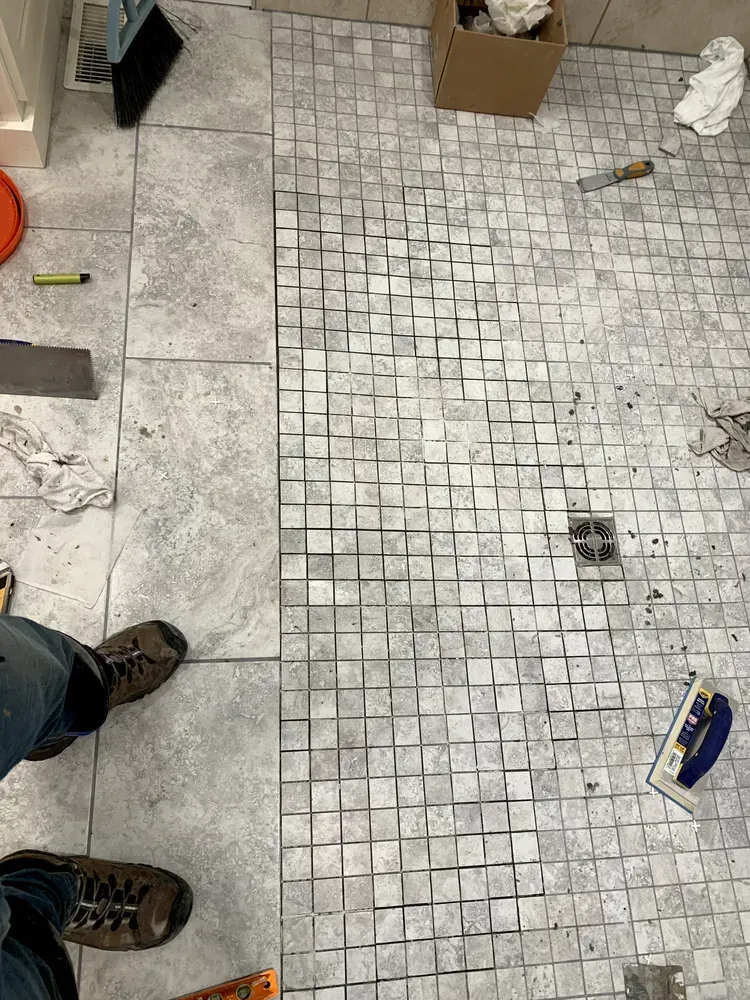 Tiled bathroom floor partially complete, with large gray tiles and mosaic tile section, tools visible.