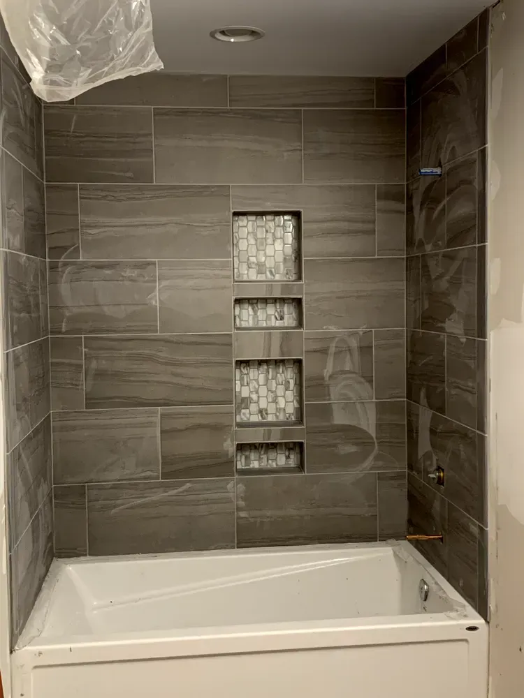Bathroom with gray tiled walls and white bathtub. Three tiled niches are filled with small mosaic tiles.