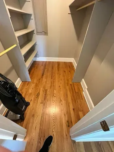 A small closet with wooden floors, built-in shelving, and hanging rod. A vacuum cleaner is in the corner.