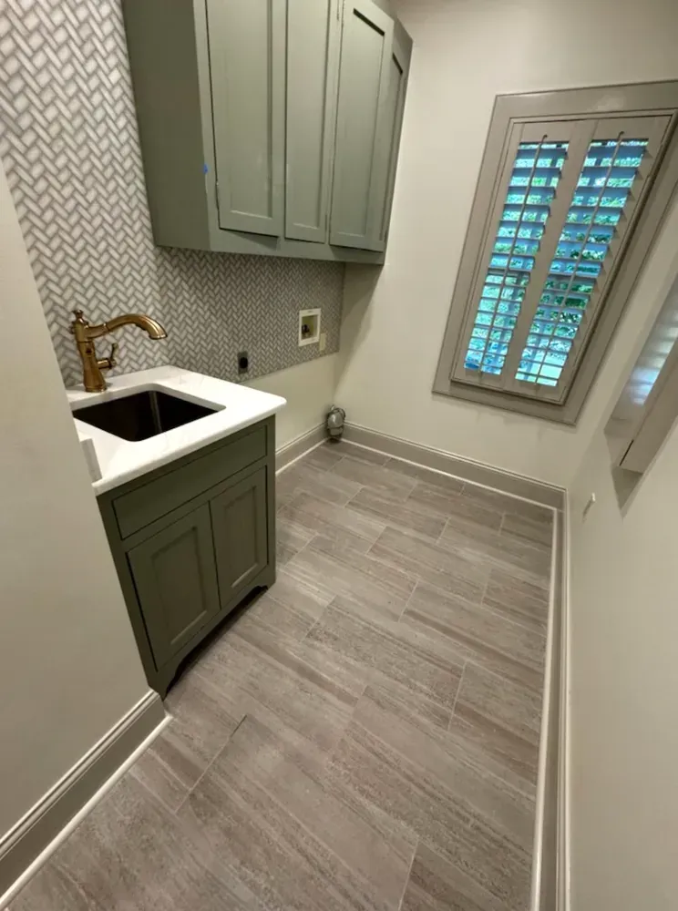 Laundry room with green cabinets, white countertop, gold faucet, and wooden floor tiles. Window with shutters.