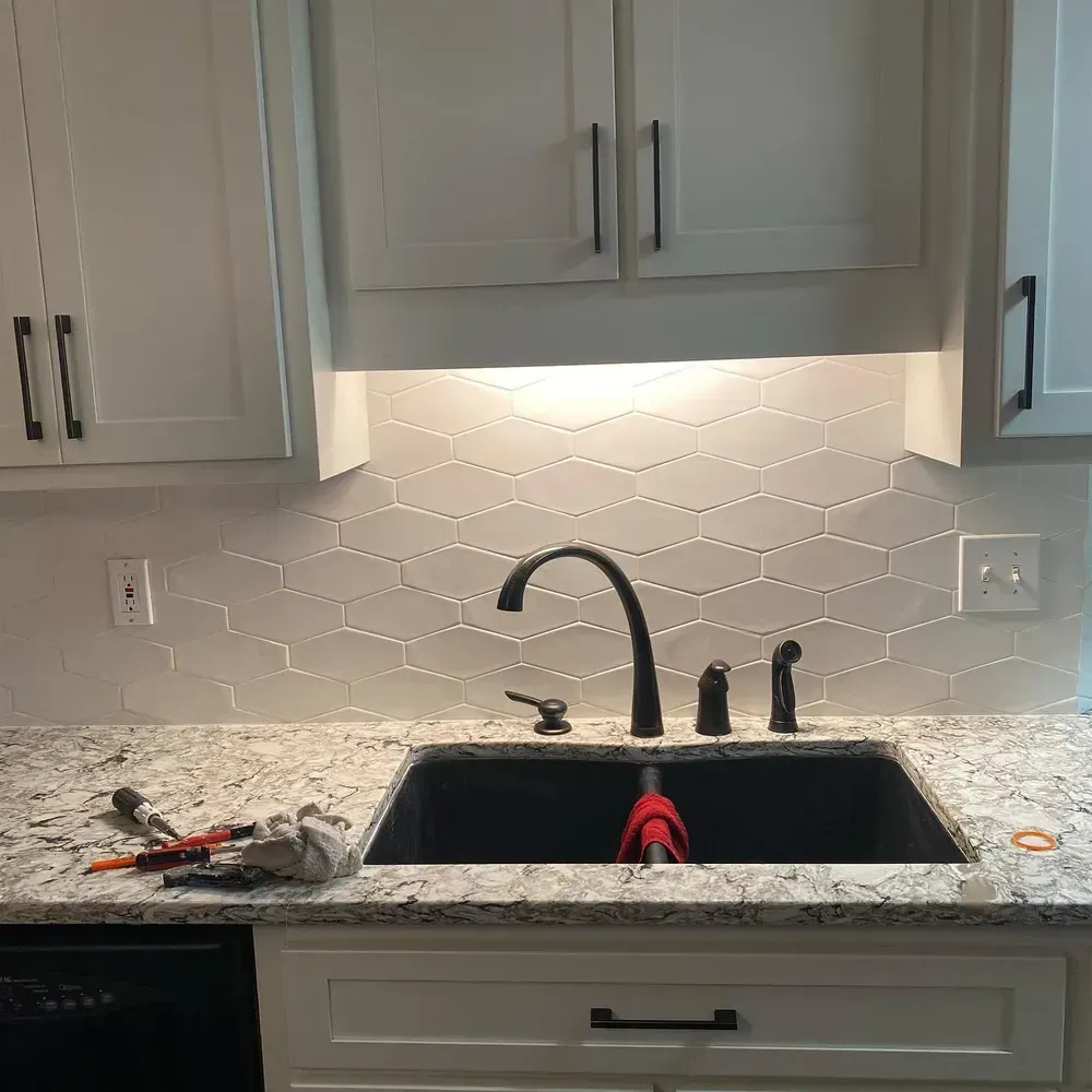 Kitchen sink with black faucet, cabinets, and granite countertop; white hexagonal tile backsplash.