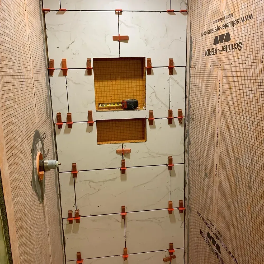 Shower wall in progress, large white tiles with orange tile leveling clips. Orange waterproof membrane visible.