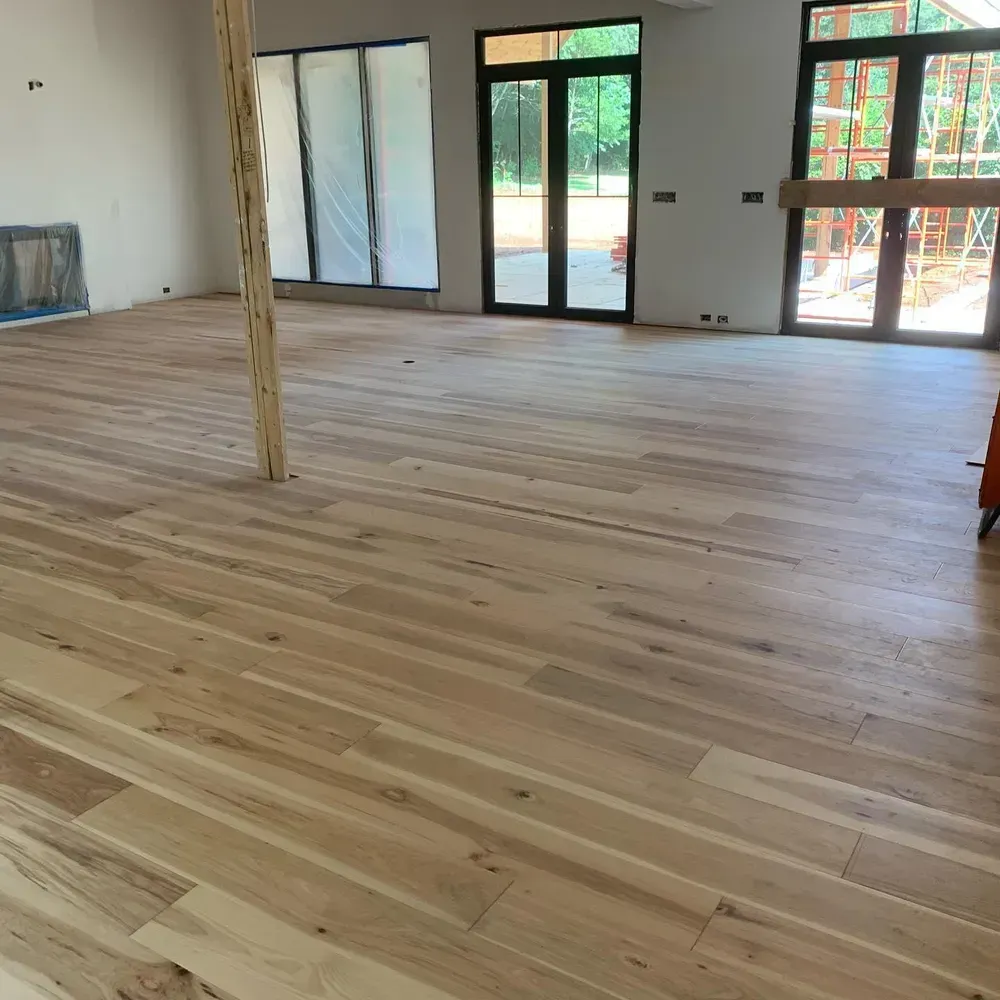 Light wood flooring installed in a room with large windows, unfinished walls, and a wooden support beam.
