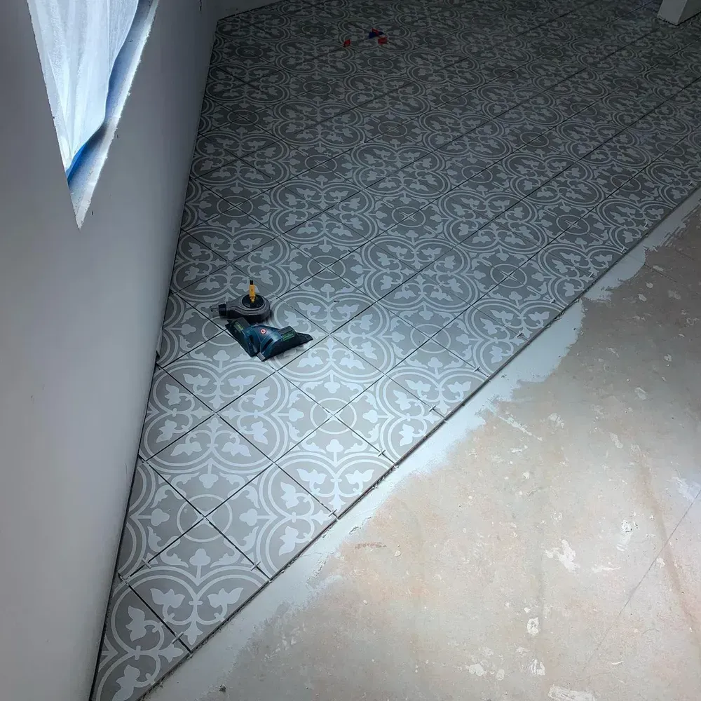 Gray patterned floor tiles being installed in a room with a white wall and concrete floor.