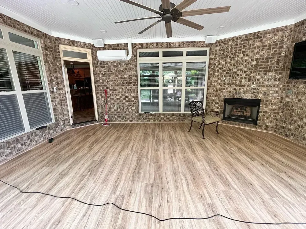 Enclosed patio with wood-look flooring, brick walls, fireplace, ceiling fan, and large windows.