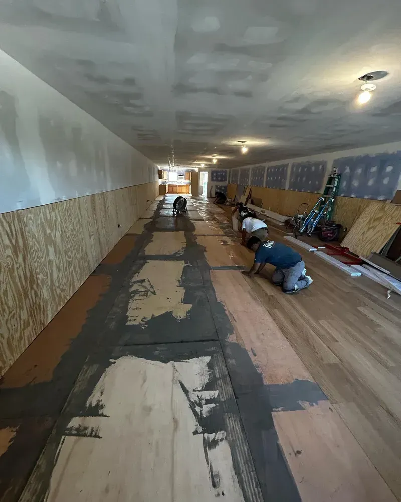 Workers installing flooring in a long, rectangular room. Walls are partially covered with plywood and drywall.