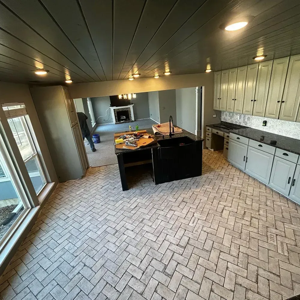 Kitchen with gray cabinets, black island, brick-patterned floor, opening to a living room with fireplace.