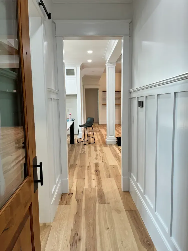 Hallway with white paneled walls, wood floors, and view into another room.