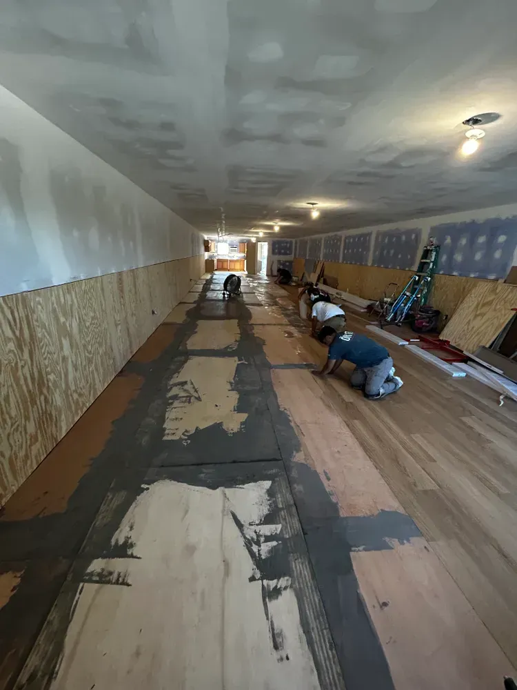 Workers installing hardwood flooring in a long, narrow room. Grey adhesive is applied to some boards.