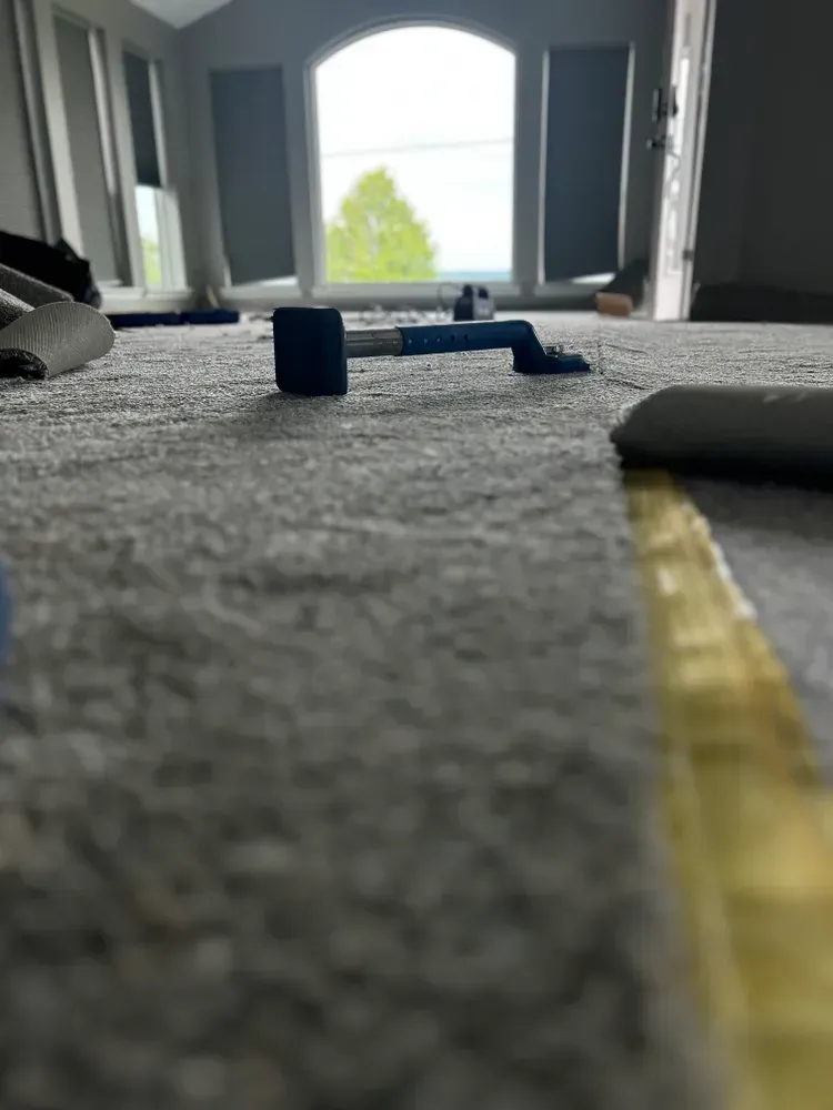 Carpet being installed in a room. Tools visible: blue hammer and yellow tape. Window in the background.