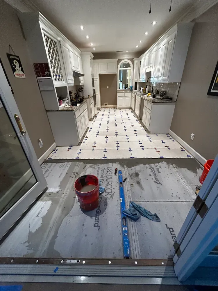 Kitchen undergoing floor tile installation; gray flooring, white cabinets. Bucket and tools in foreground.