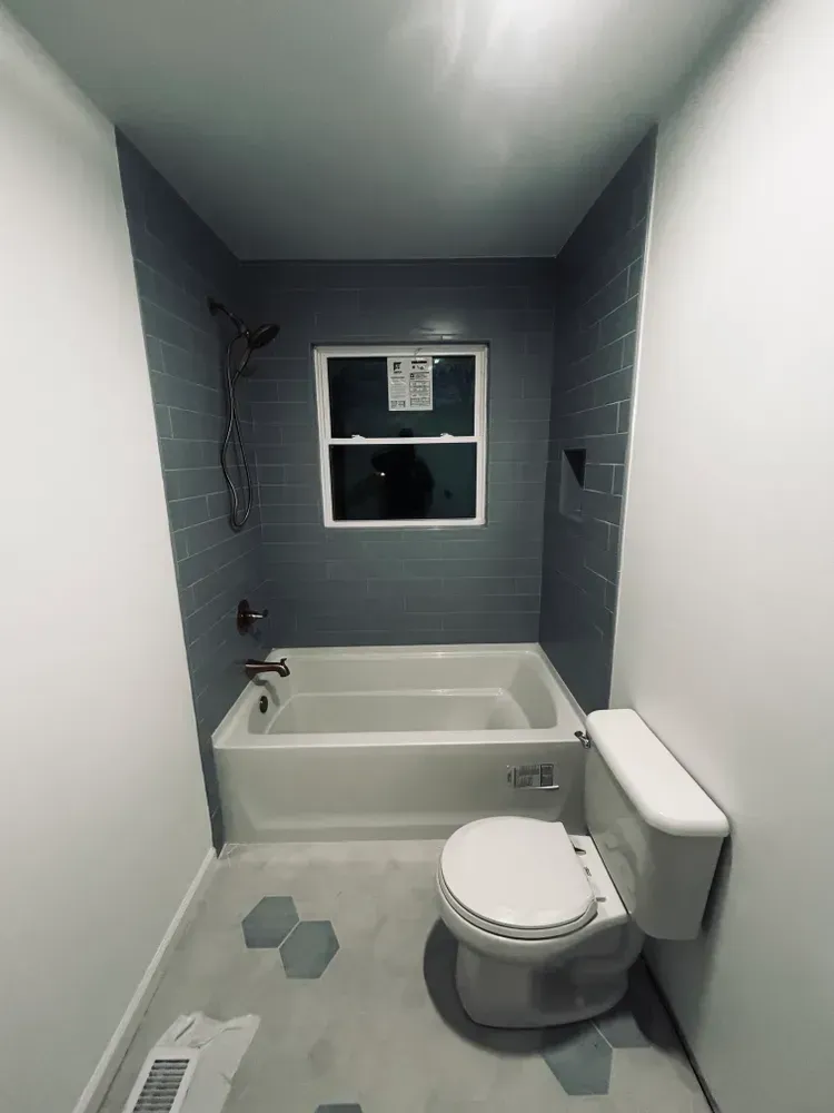 Modern bathroom with gray tiled shower wall, white tub and toilet, gray and white hexagon floor tiles, and a window.