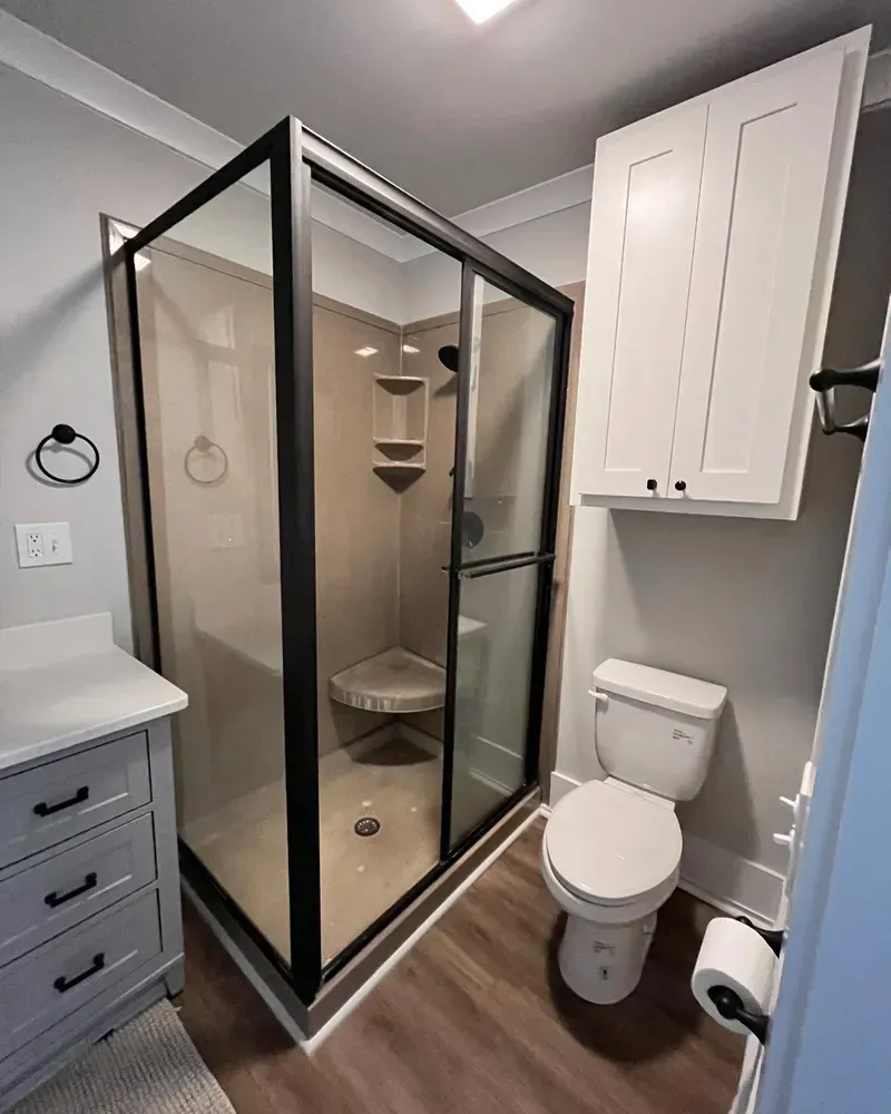 Bathroom with a black-framed glass shower, toilet, and white cabinets against light gray walls and brown flooring.