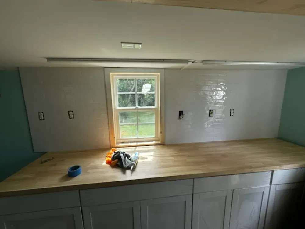 Kitchen renovation: white tiled backsplash, wooden countertop, window, white cabinets, and tools on countertop.