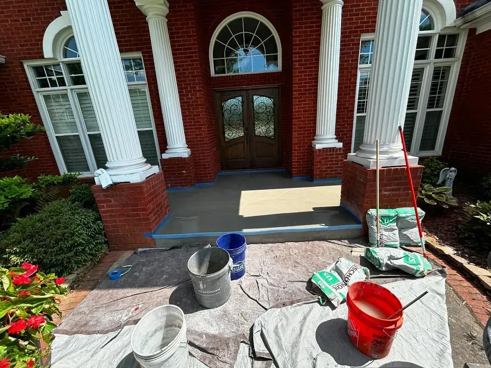 Front porch being resurfaced, buckets, tools, and materials are visible. Red brick, white columns, and brown door.