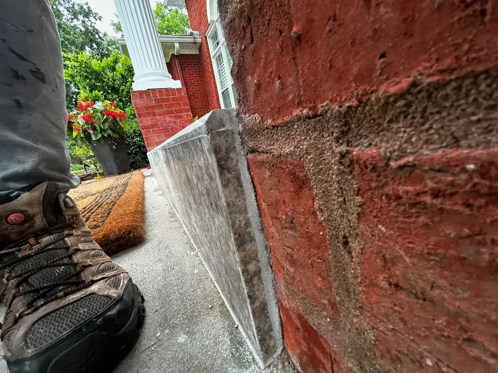 Close-up of brick wall and concrete steps next to a person's boot and a welcome mat outside a building.