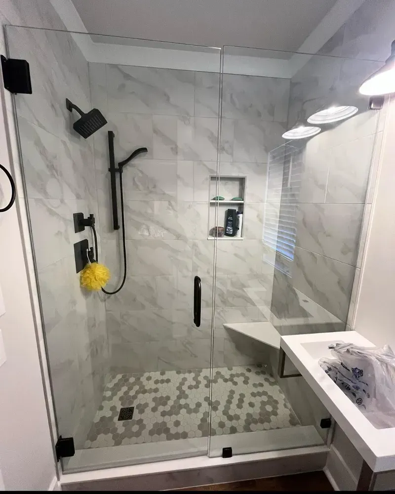 Modern shower with gray marble-look tile, black fixtures, glass door, built-in seat, and hexagon floor tile.
