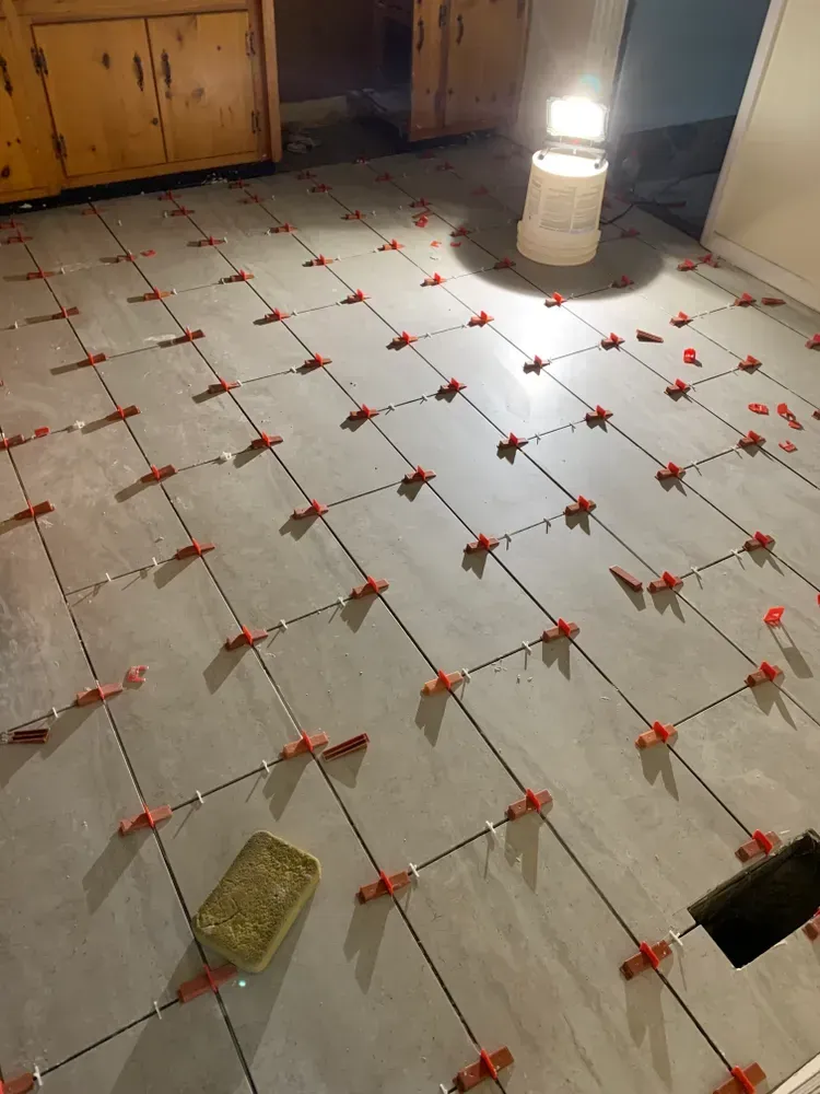 Gray tiles on a floor, red leveling clips in use. A lit work light sits in the background.
