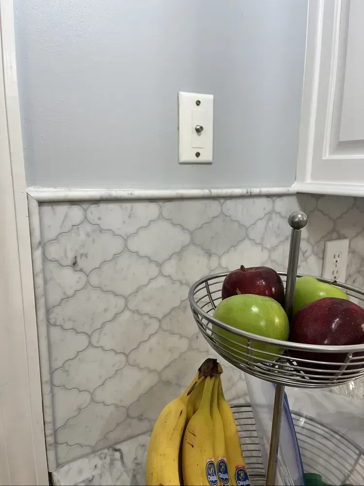 White wall with marble backsplash, fruit basket, and a phone jack. Bananas and apples in view.