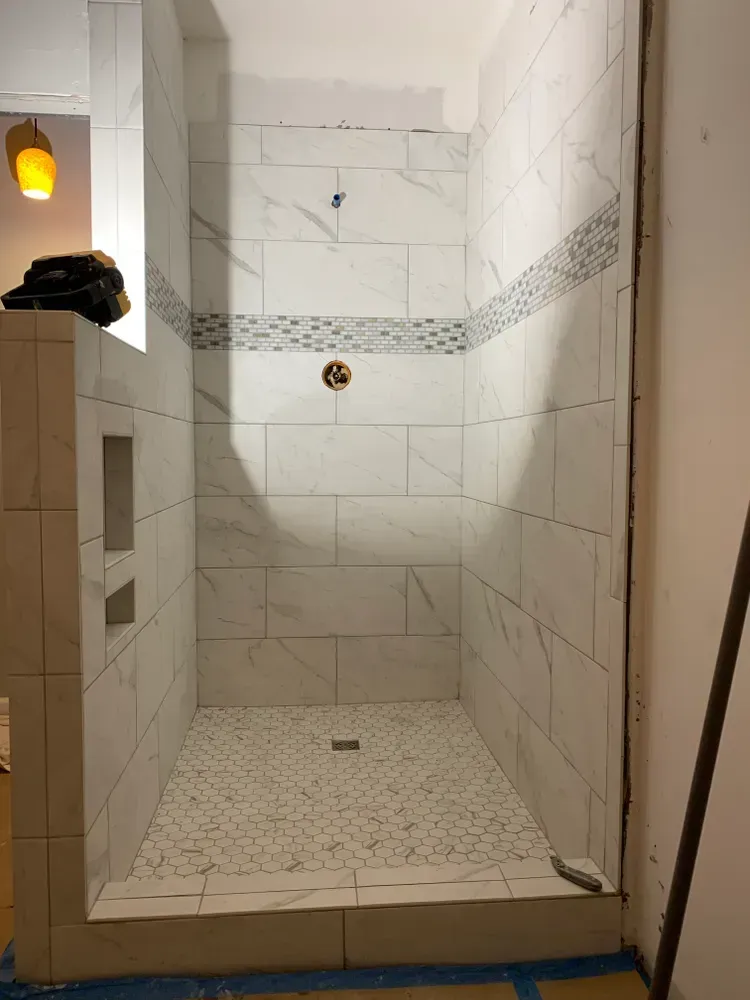 Tiled shower stall under construction with white marble-look tiles, mosaic accent band, and pebble floor.