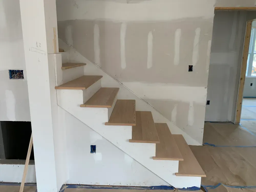 Staircase with light wood treads and white risers, in a room under construction, with drywall walls.