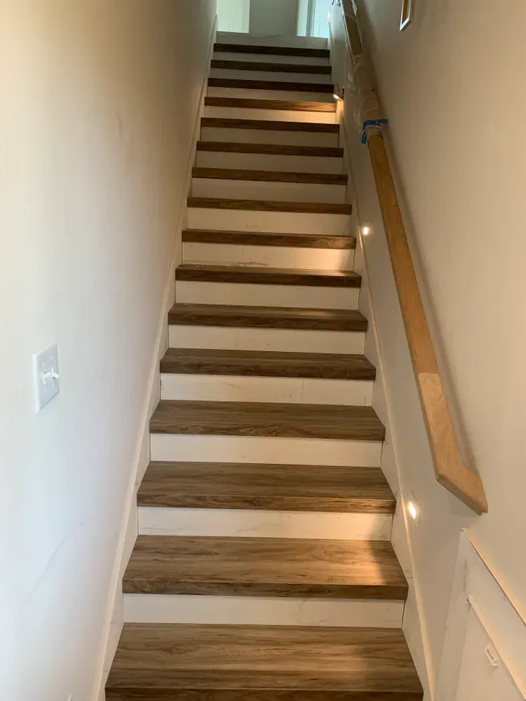 Staircase with wood-look treads, white risers, and a wooden handrail on the right. Stairwell is lit from above and recessed lighting.