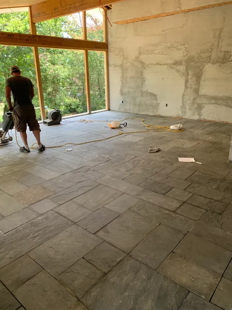 Interior of a room under construction with gray stone tile flooring. A person stands near tools, and a large window looks out to trees.