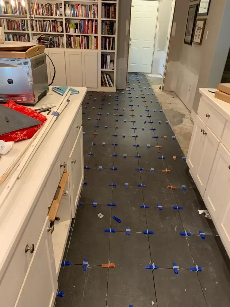Kitchen floor tile installation in progress; black tiles with levelers. White cabinets, bookshelf in the background.