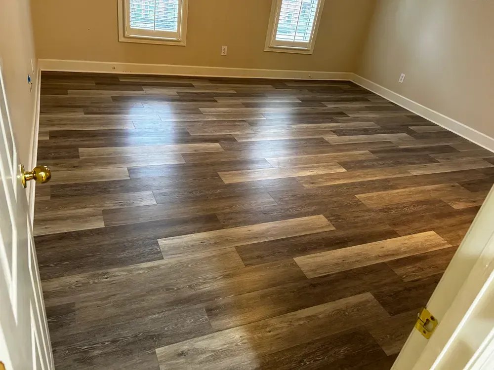 Empty room with new wood-look flooring, two windows with white trim, tan walls and white baseboards.