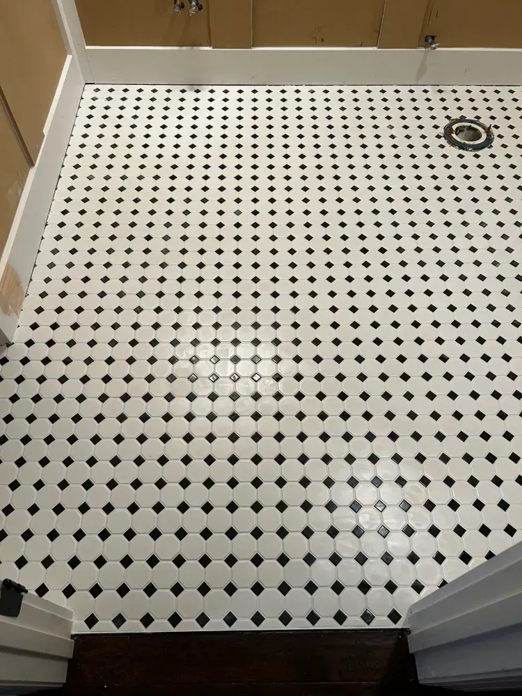 Black and white tiled floor with a drain, viewed from doorway, under wooden cabinets.
