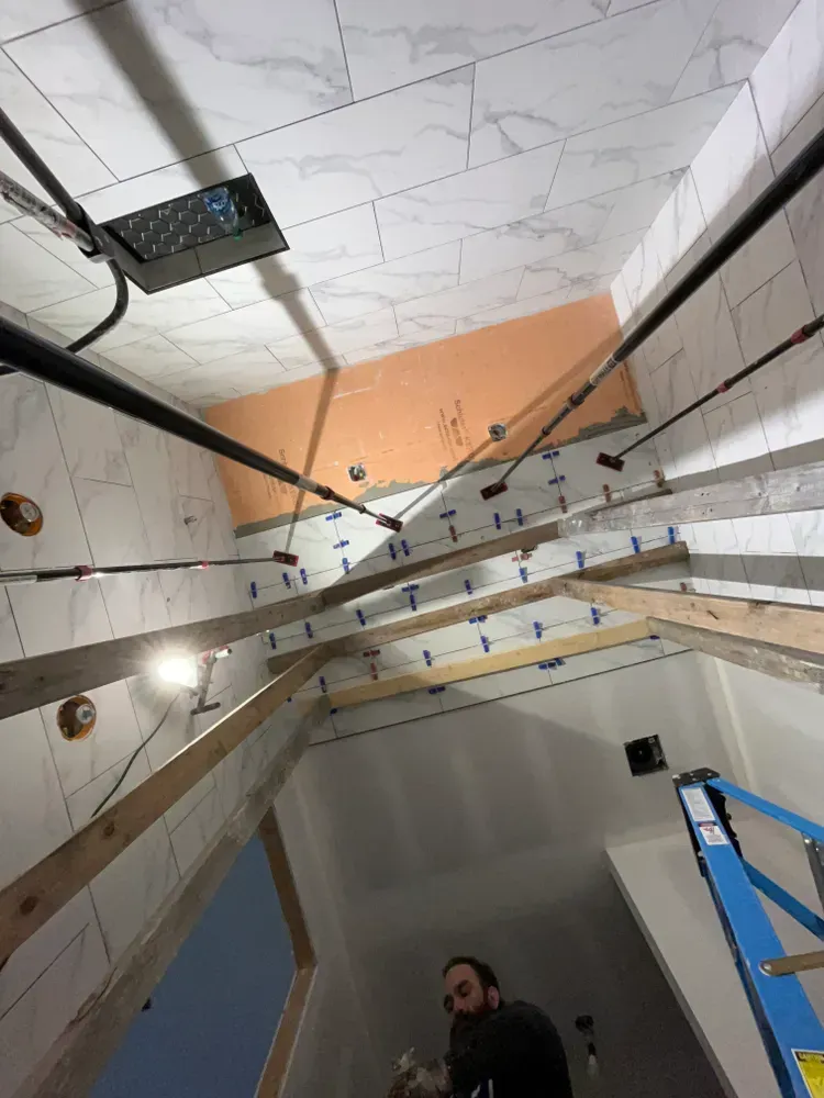 A construction worker installs tiles in a bathroom, with wood beams and pipes visible.