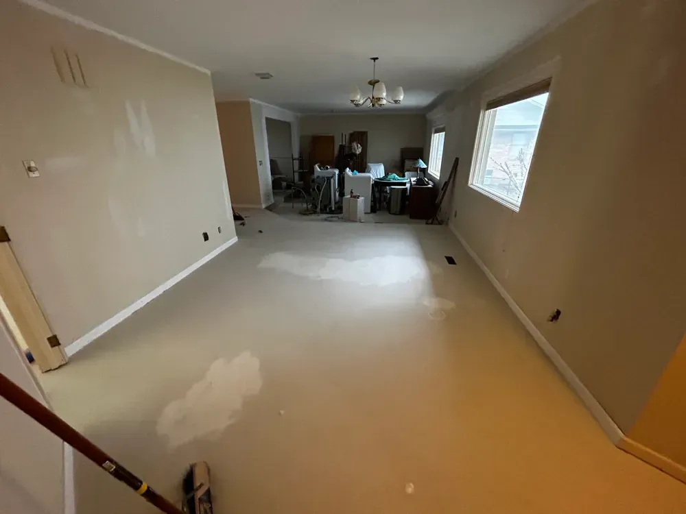 Room with tan carpet and walls, partially painted, with miscellaneous items in the background.