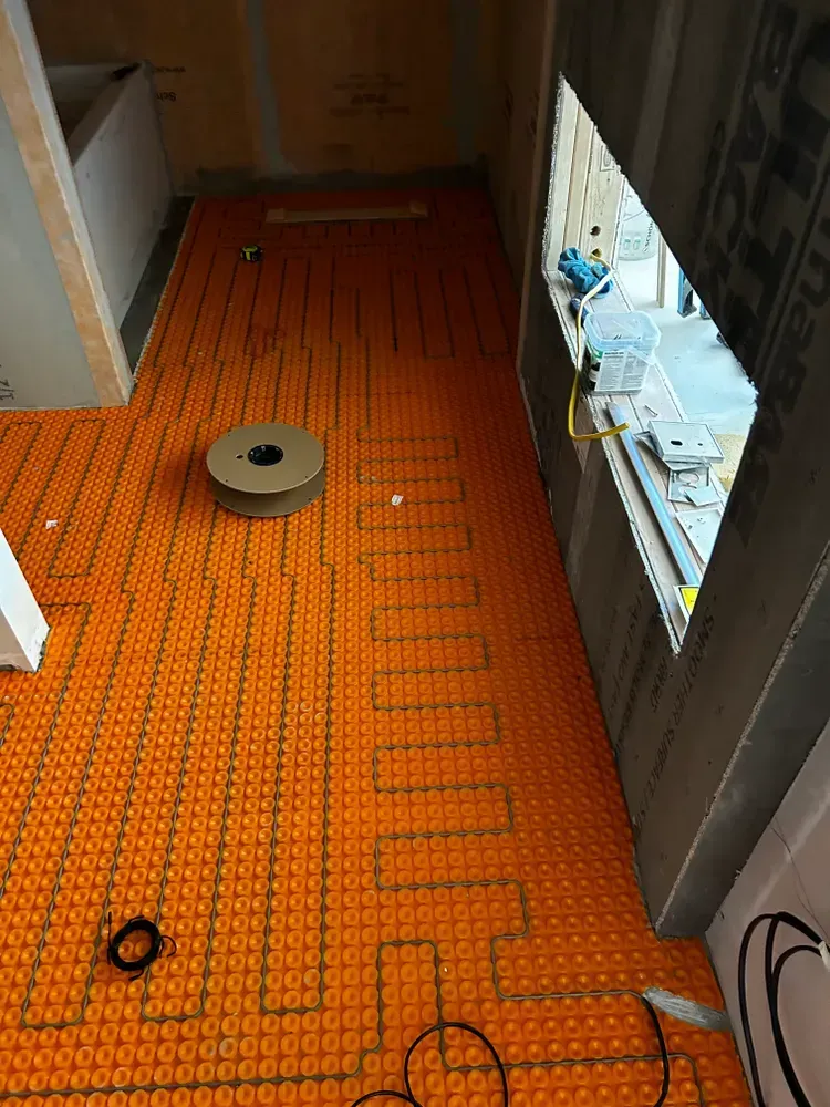 Orange heated floor installation in a room with a window, roll of cable, and unfinished walls.