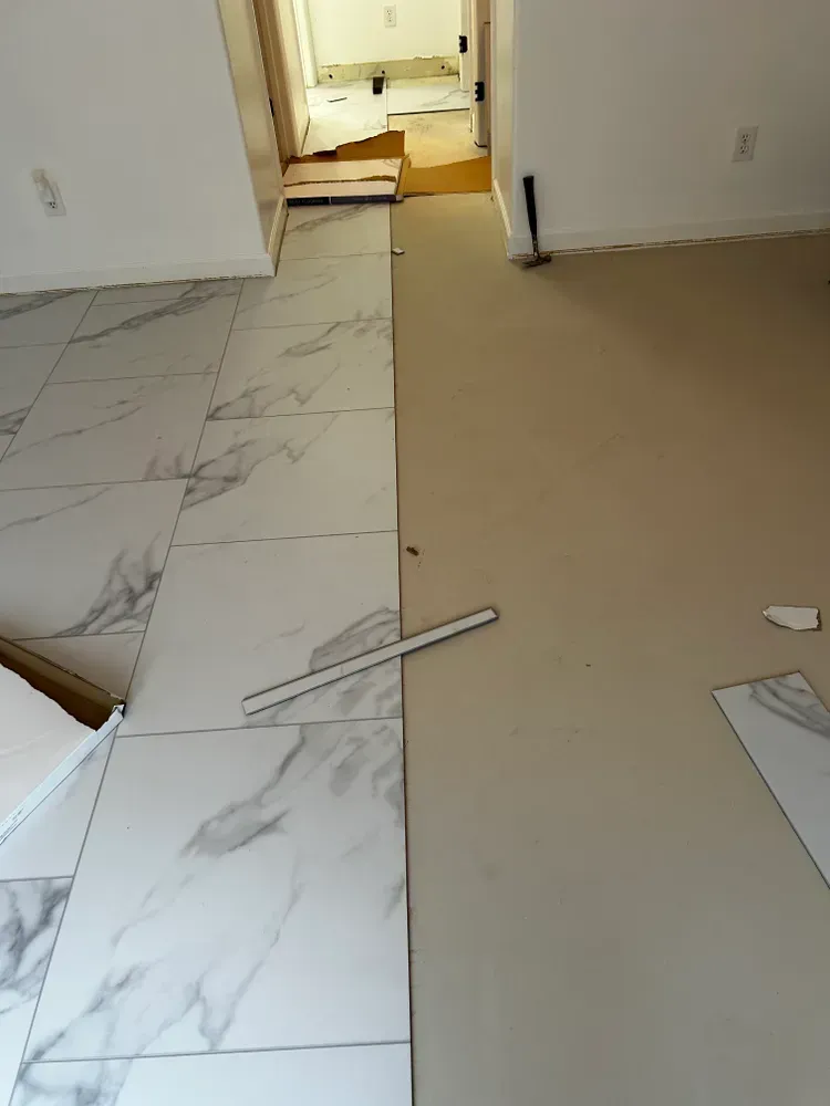 White marble-look tiles installed on one side of a room, unfinished floor on the other.