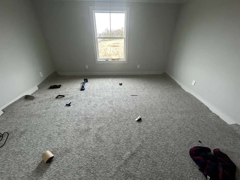 Empty room with gray carpet, walls, and window. Debris scattered on the floor.