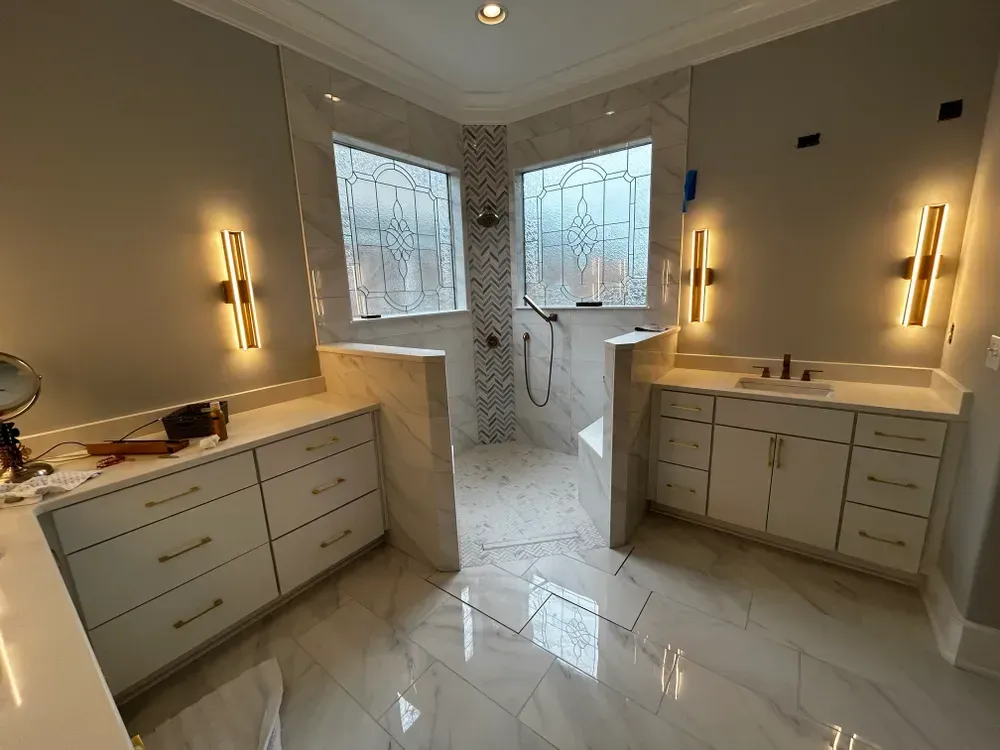 Modern bathroom with white cabinets, marble floors, and glass shower. Gold sconces on walls.